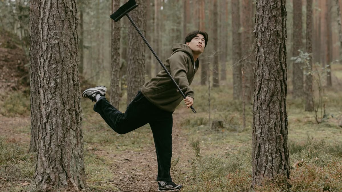 A man playfully balances a broomstick in a lush forest, evoking creativity.