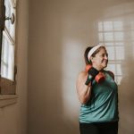 Senior woman in a brightly lit room exercises with dumbbells, promoting fitness at home.