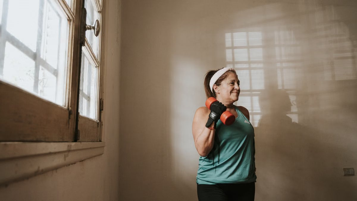 Senior woman in a brightly lit room exercises with dumbbells, promoting fitness at home.