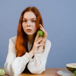 Woman with red hair holding lime surrounded by fresh produce on table.