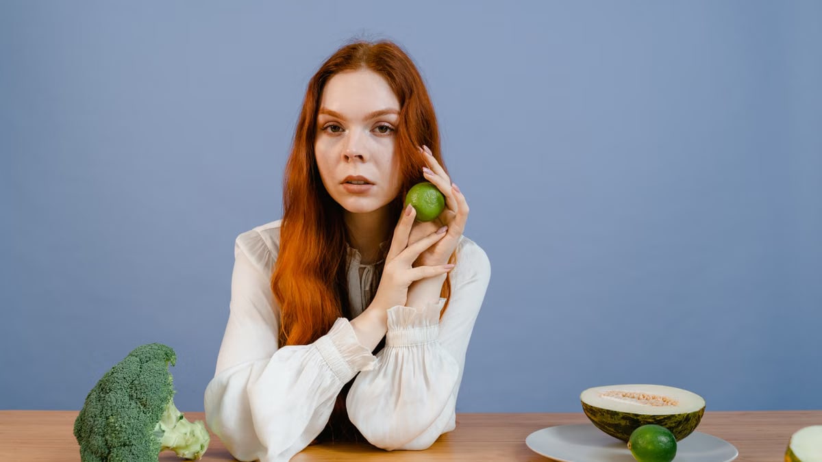 Woman with red hair holding lime surrounded by fresh produce on table.